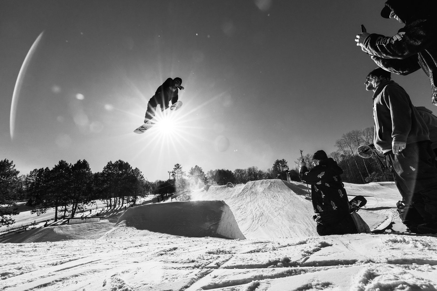 Snowboarder performs aerial trick over snow jump at sunny ski park in black and white photo.