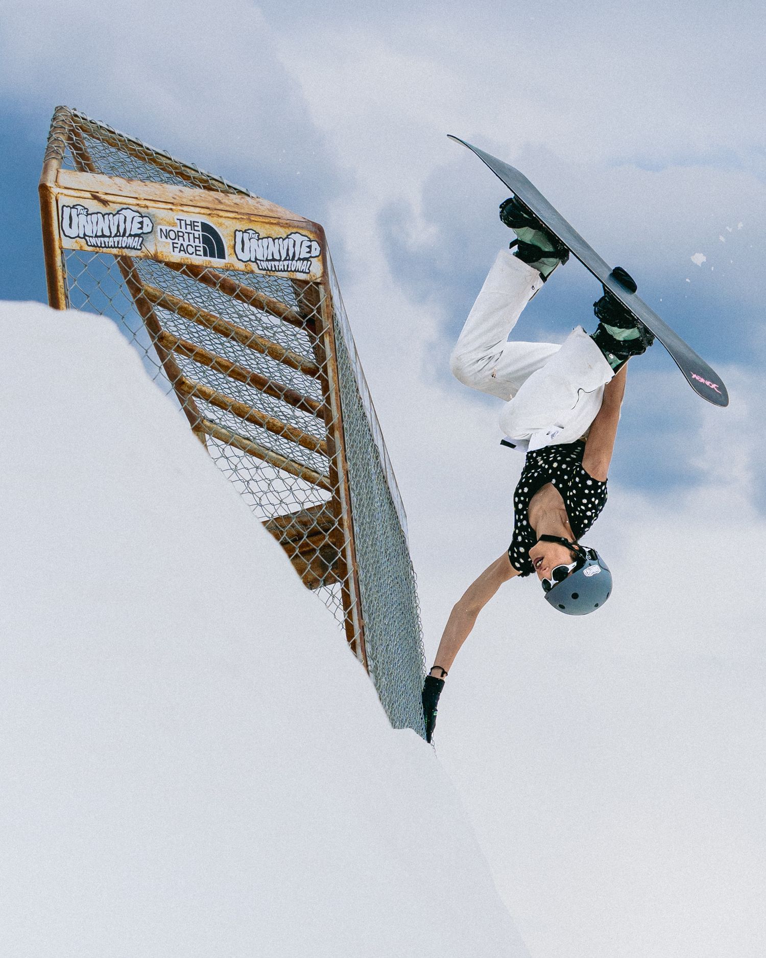 Snowboarder performs an inverted aerial trick at a snow park competition near a chain-link fence feature.