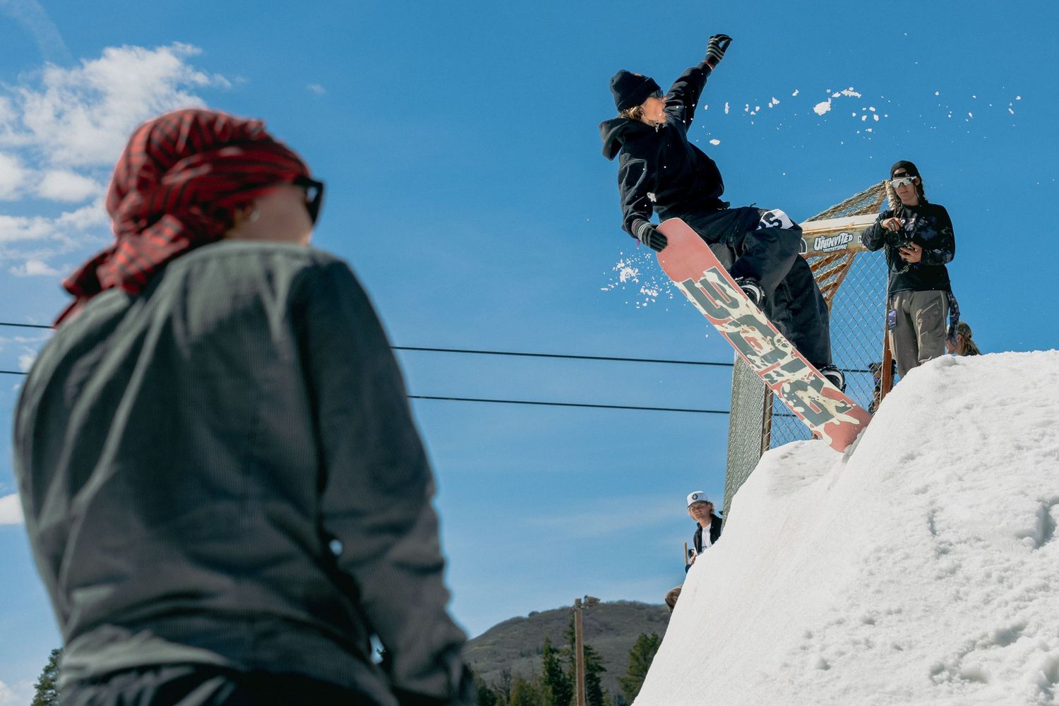 Snowboarder performs a trick on a snowy rail while spectators watch under a bright blue sky at a winter terrain park.