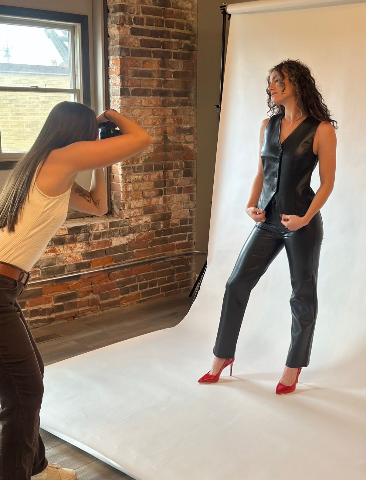 A person in black attire and red heels poses against a white backdrop and exposed brick wall in a studio setting.