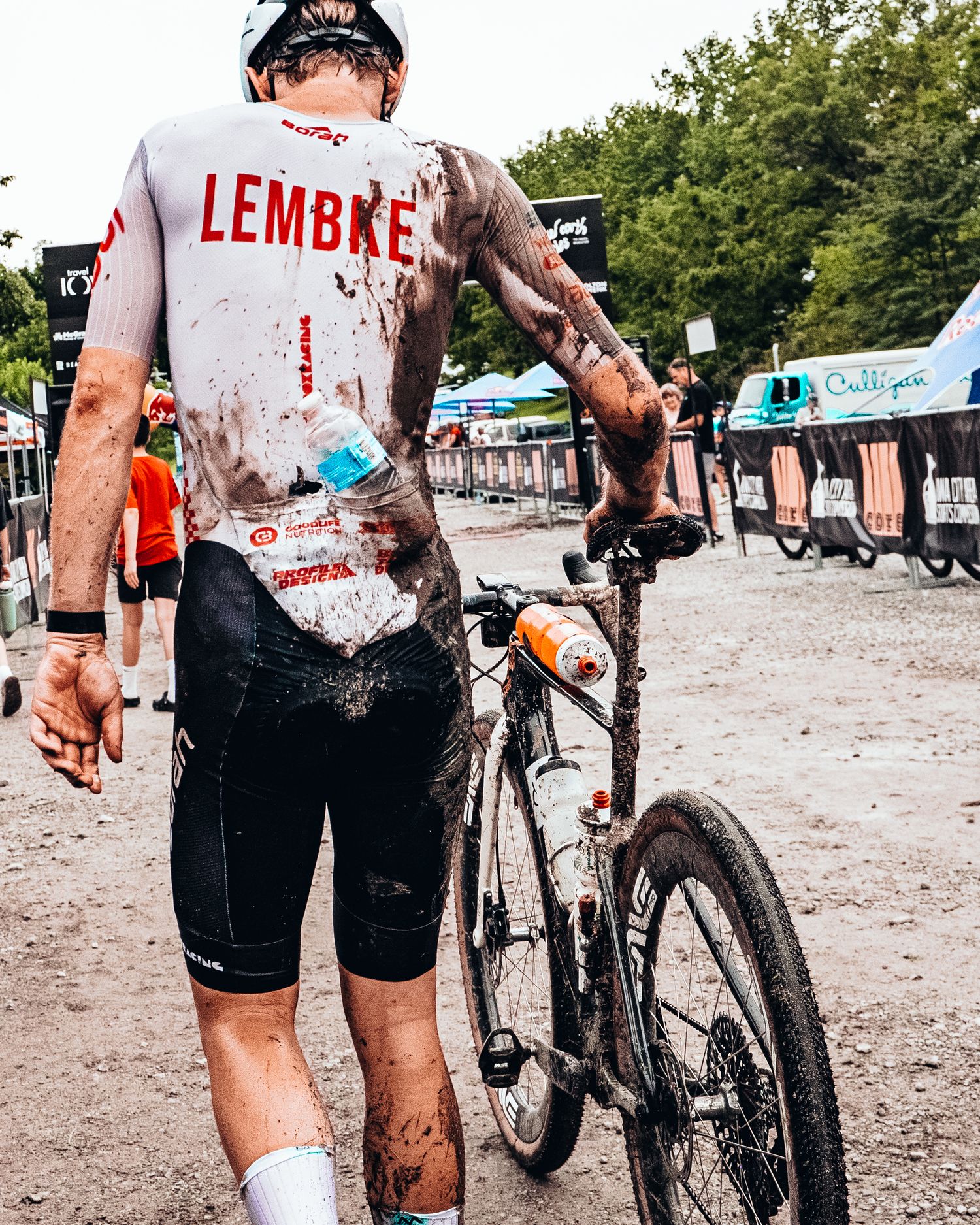 Muddy cyclist named Lembke walks bike after a grueling gravel race, covered in dirt at outdoor event.