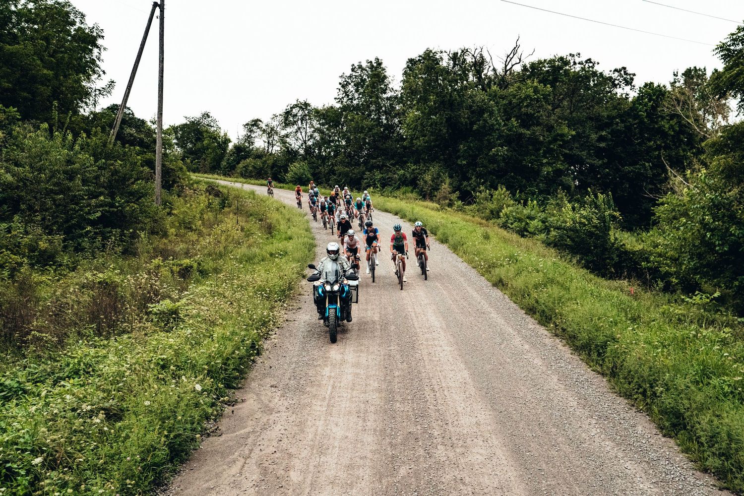 A group of cyclists race down a rural gravel road flanked by lush green trees, led by a motorcycle pace vehicle.