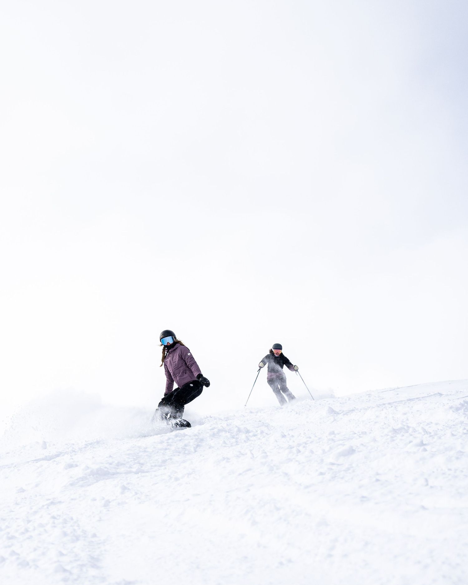 Two skiers descending down a snow-covered slope against white winter sky.