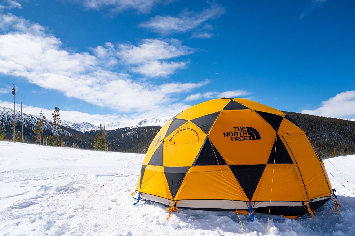 Bright yellow geometric tent pitched in snow with mountain peaks in background.