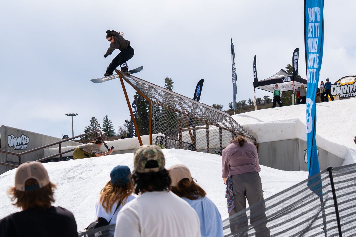 Snowboarder performing a rail grind trick at an outdoor winter sports competition with spectators watching.