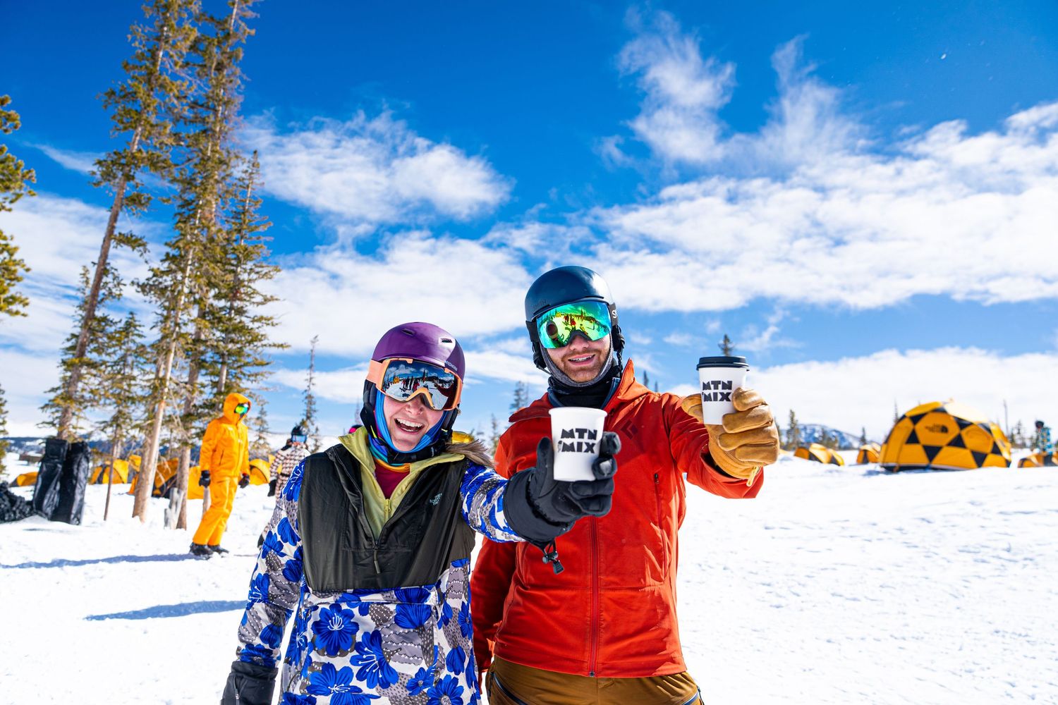 Two winter sports enthusiasts in ski gear toasting drinks in snowy mountain setting.