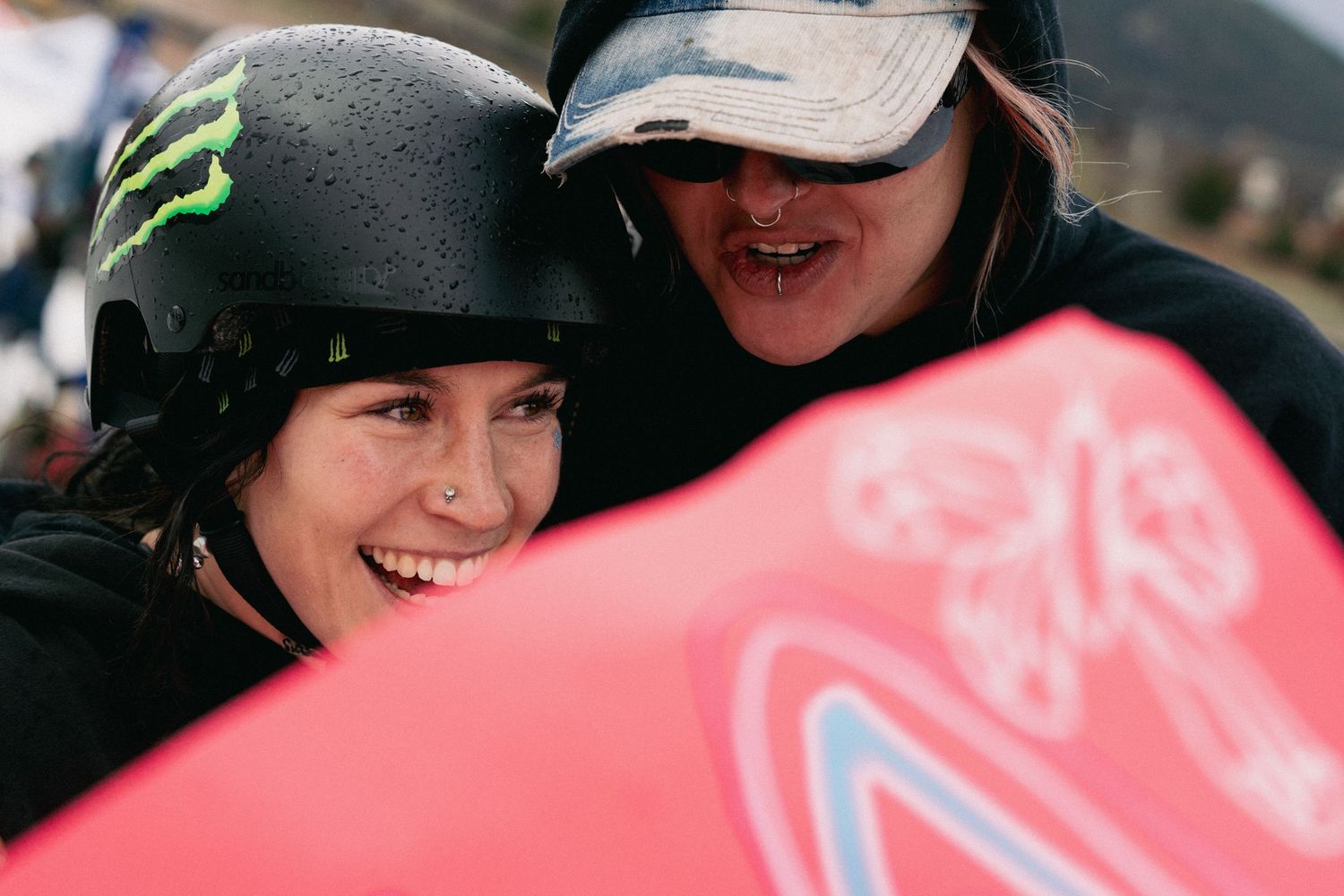 Two smiling snowboarders celebrate together, one wearing a Monster Energy helmet at a winter sports competition.