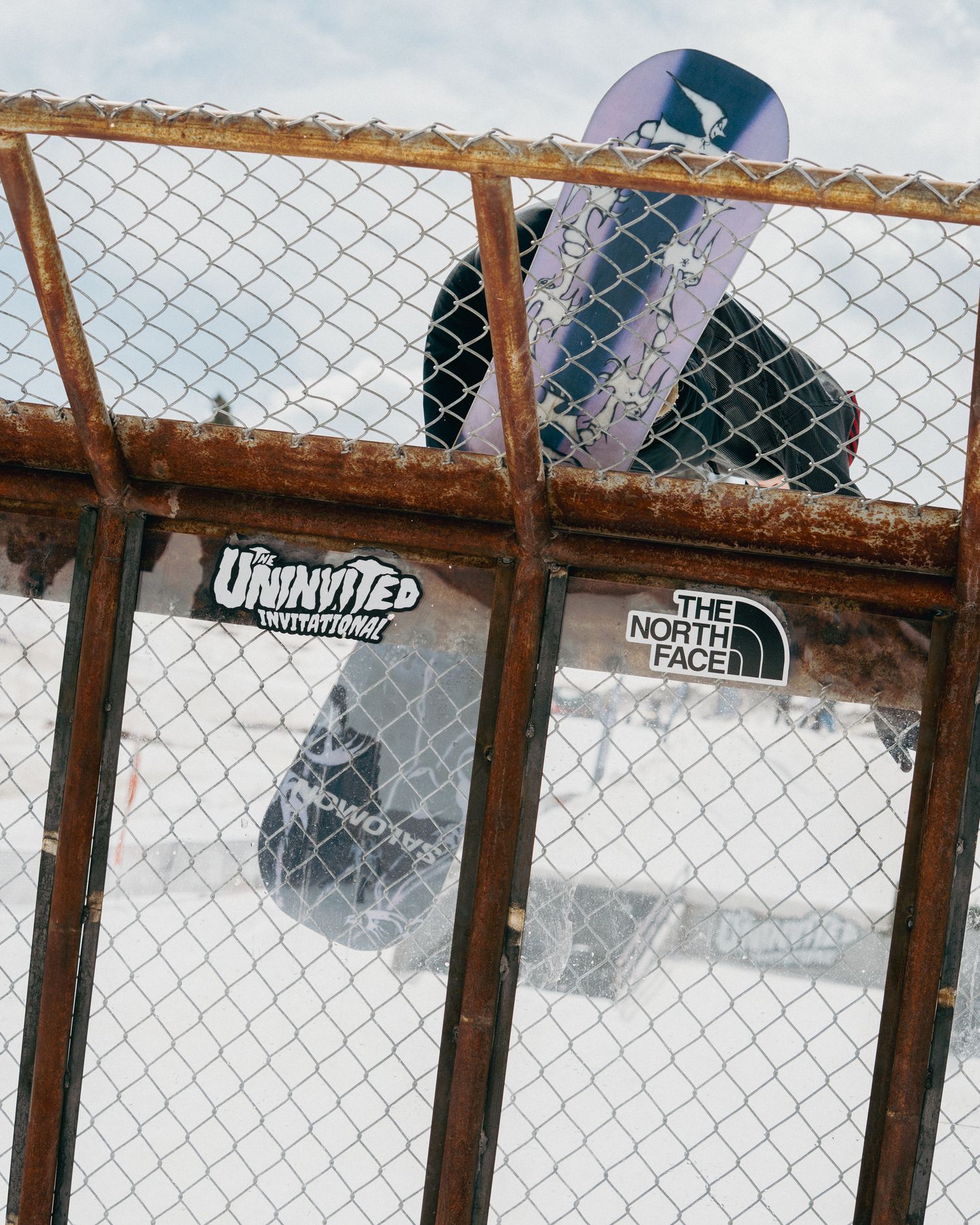 Snowboarder carrying a board behind a rusty chain-link fence with The North Face and Uninvited stickers.