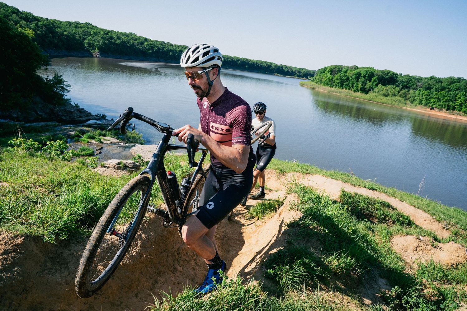 A cyclist carries a gravel bike up a steep dirt trail near a river during an outdoor cycling race.