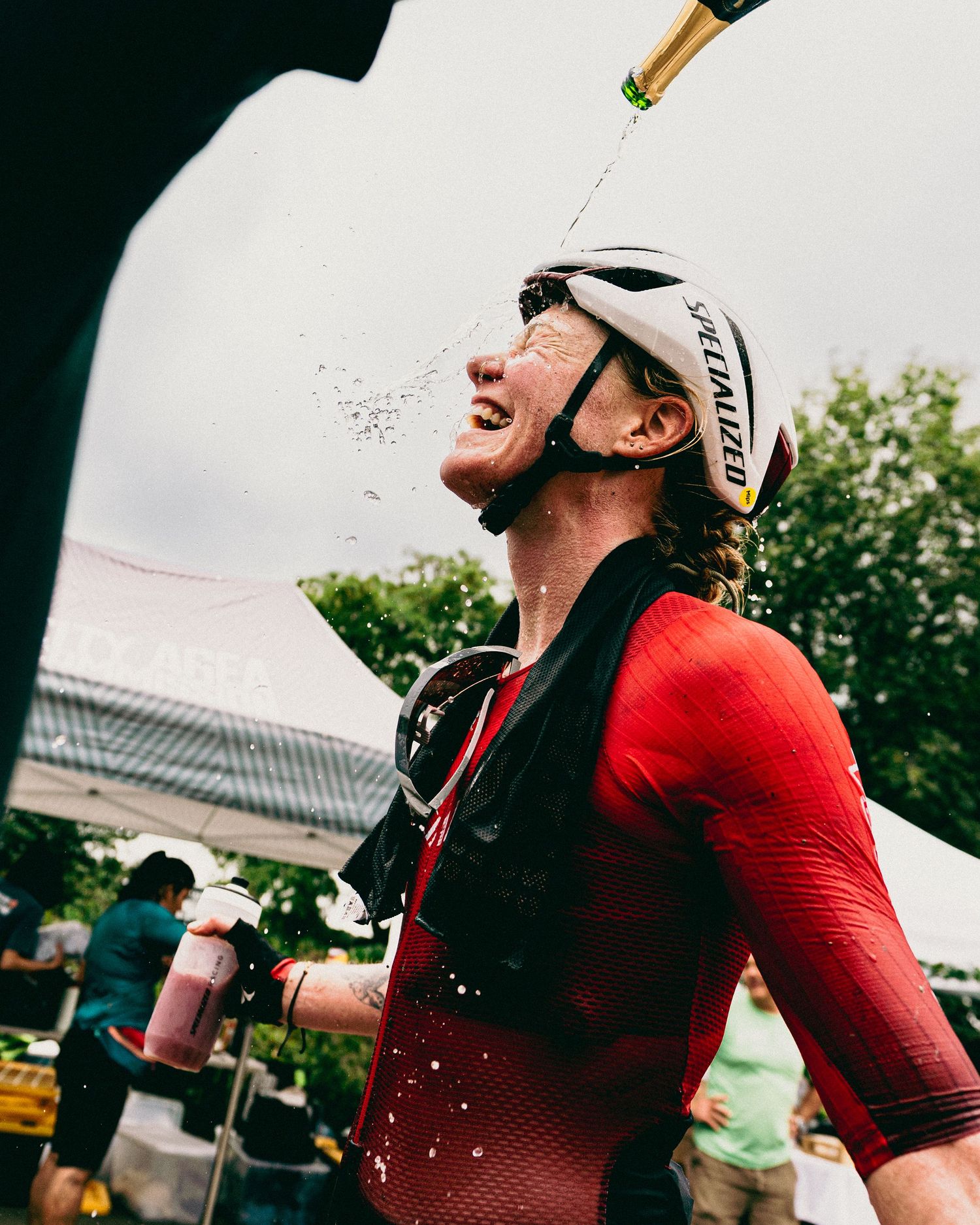 Cyclist in red jersey and Specialized helmet celebrates with champagne shower at outdoor cycling event finish line.