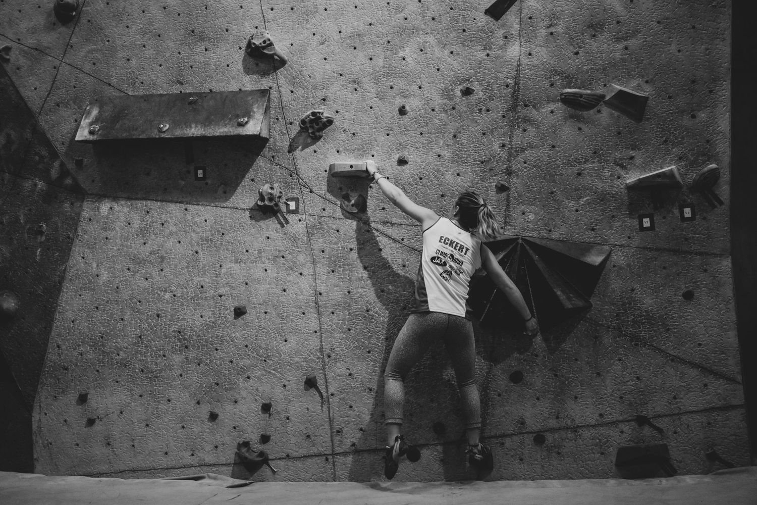 Athletic climber scales overhanging section of indoor climbing wall in dramatic black and white shot.