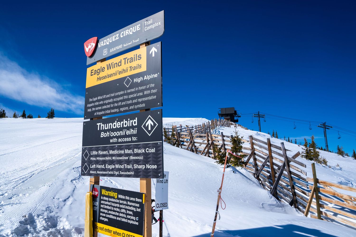 Trail information board at ski resort with wooden fence and snowy slopes in background.