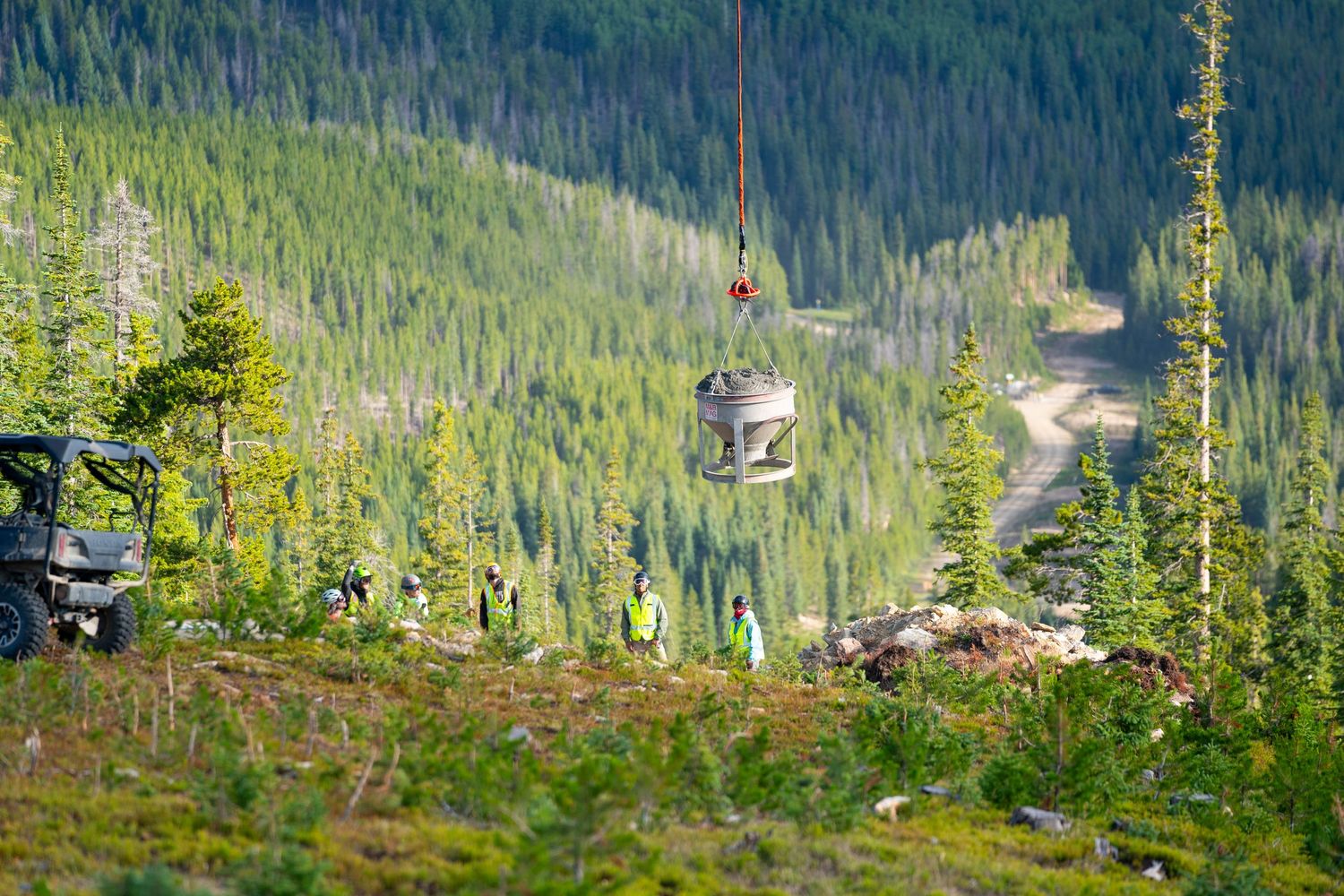 Aerial transport carrying equipment over forested mountain valley with ski infrastructure visible below.