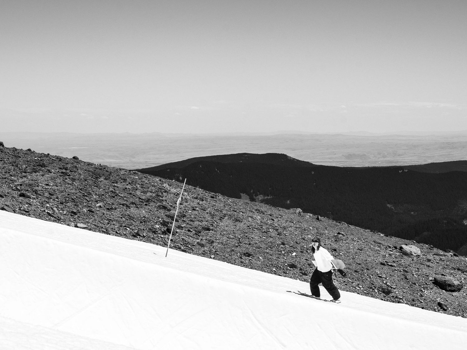 Black and white image of skier descending snowy mountain slope with distant hills visible.