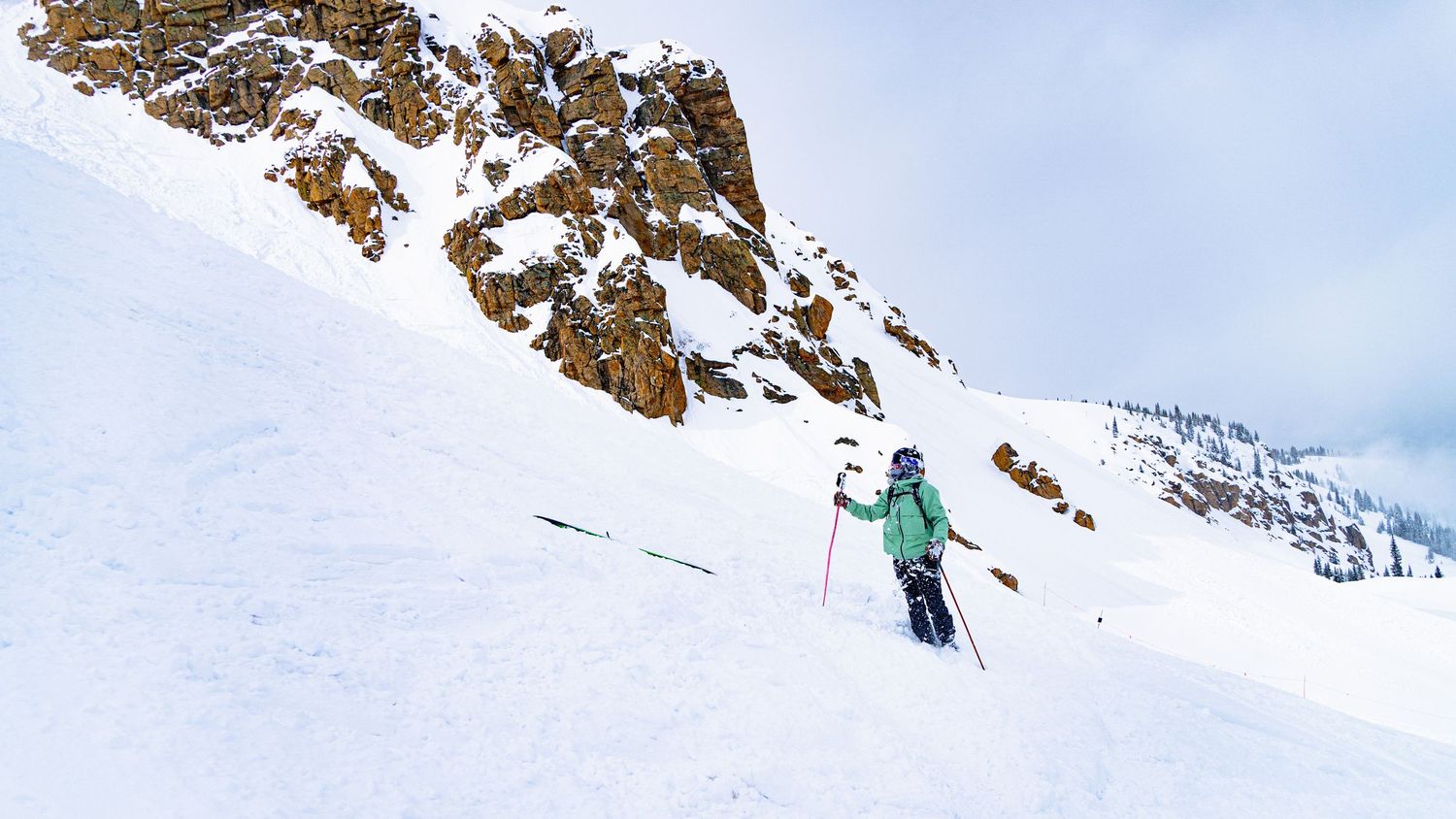 A skier descends a steep snowy slope with dramatic rocky peaks in the background.