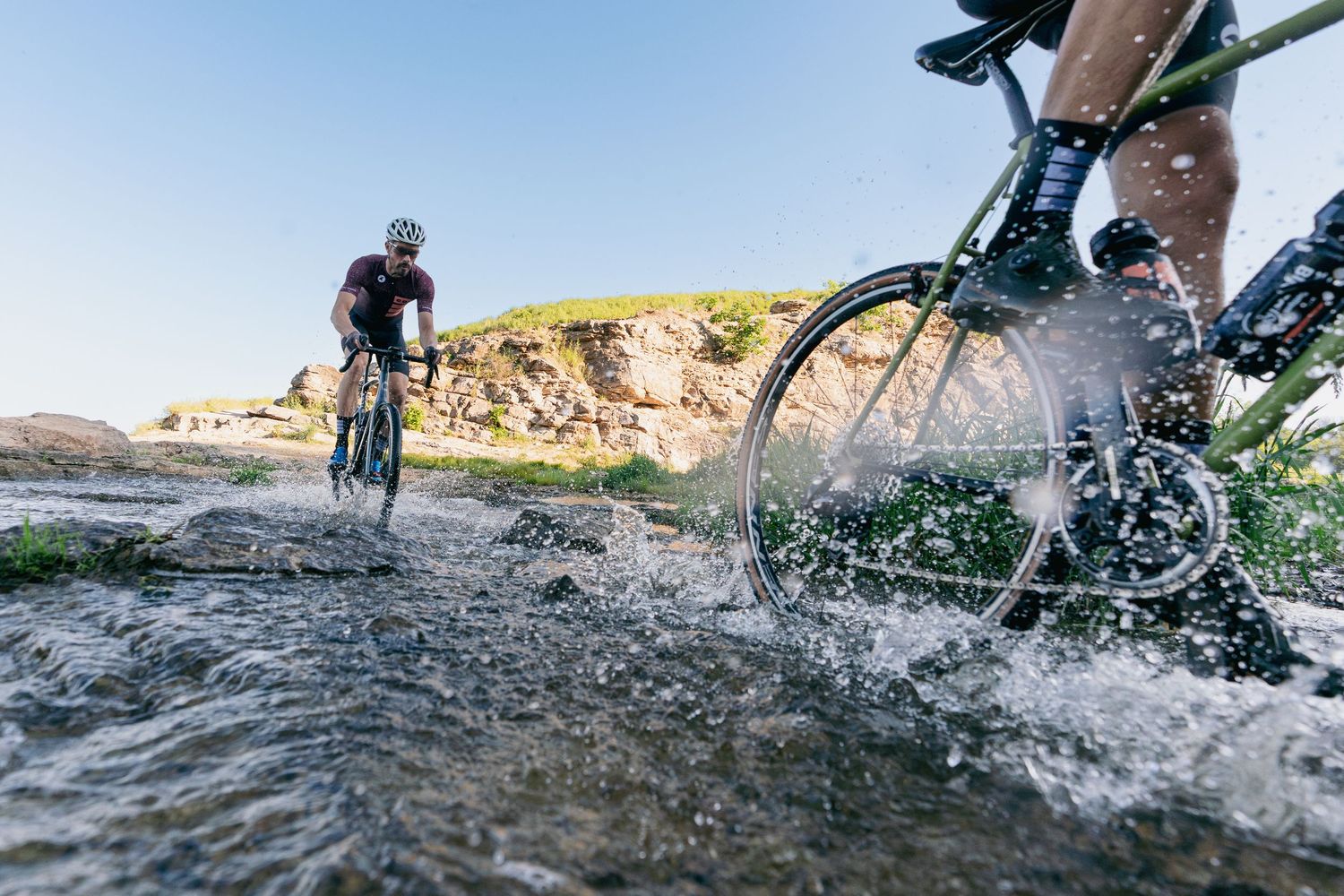 Two mountain bikers splash through a shallow river crossing on a sunny day with rocky cliffs in the background.