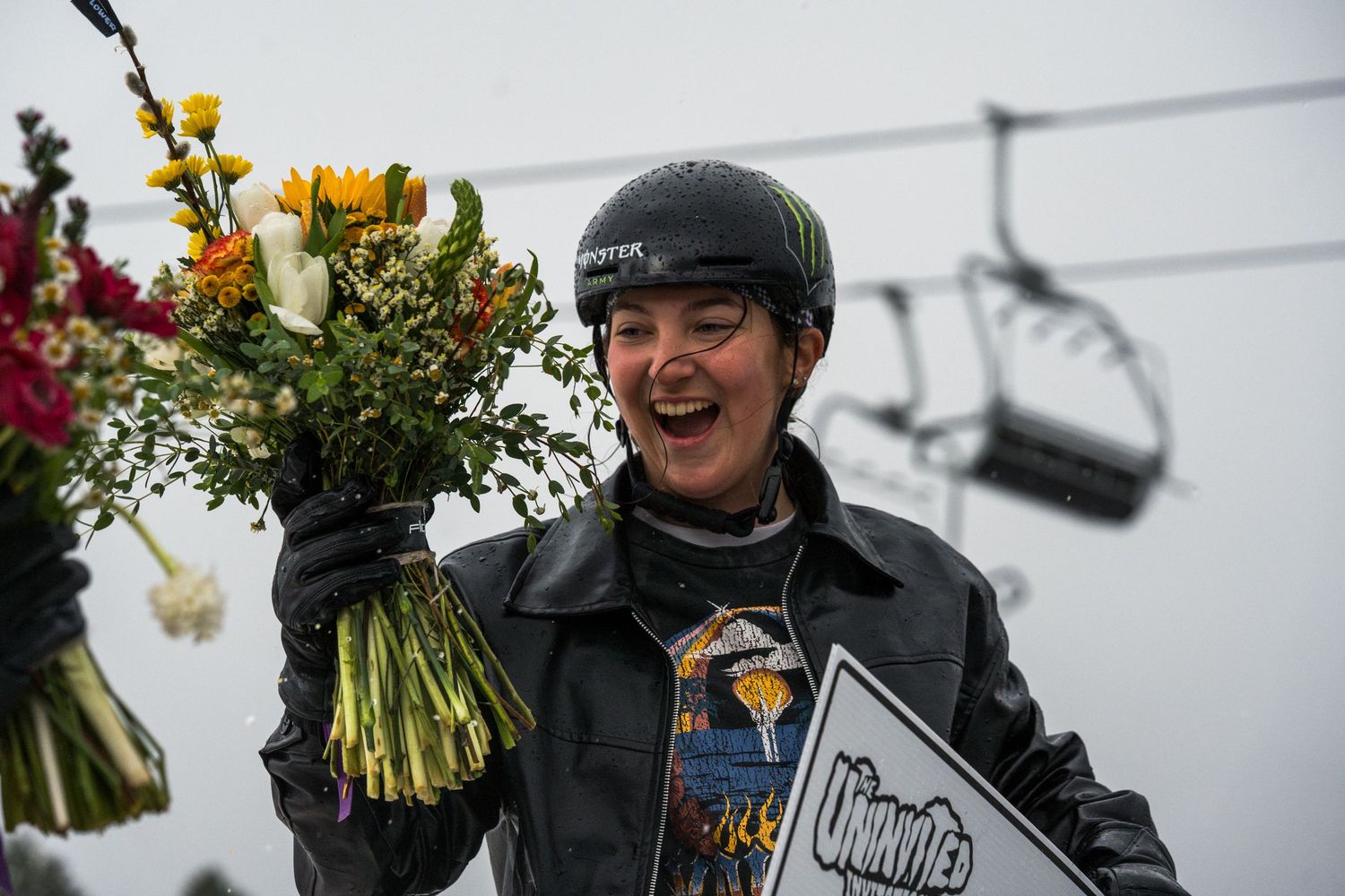 Smiling female snowboarder holding flowers and a winner's sign at a ski resort competition.