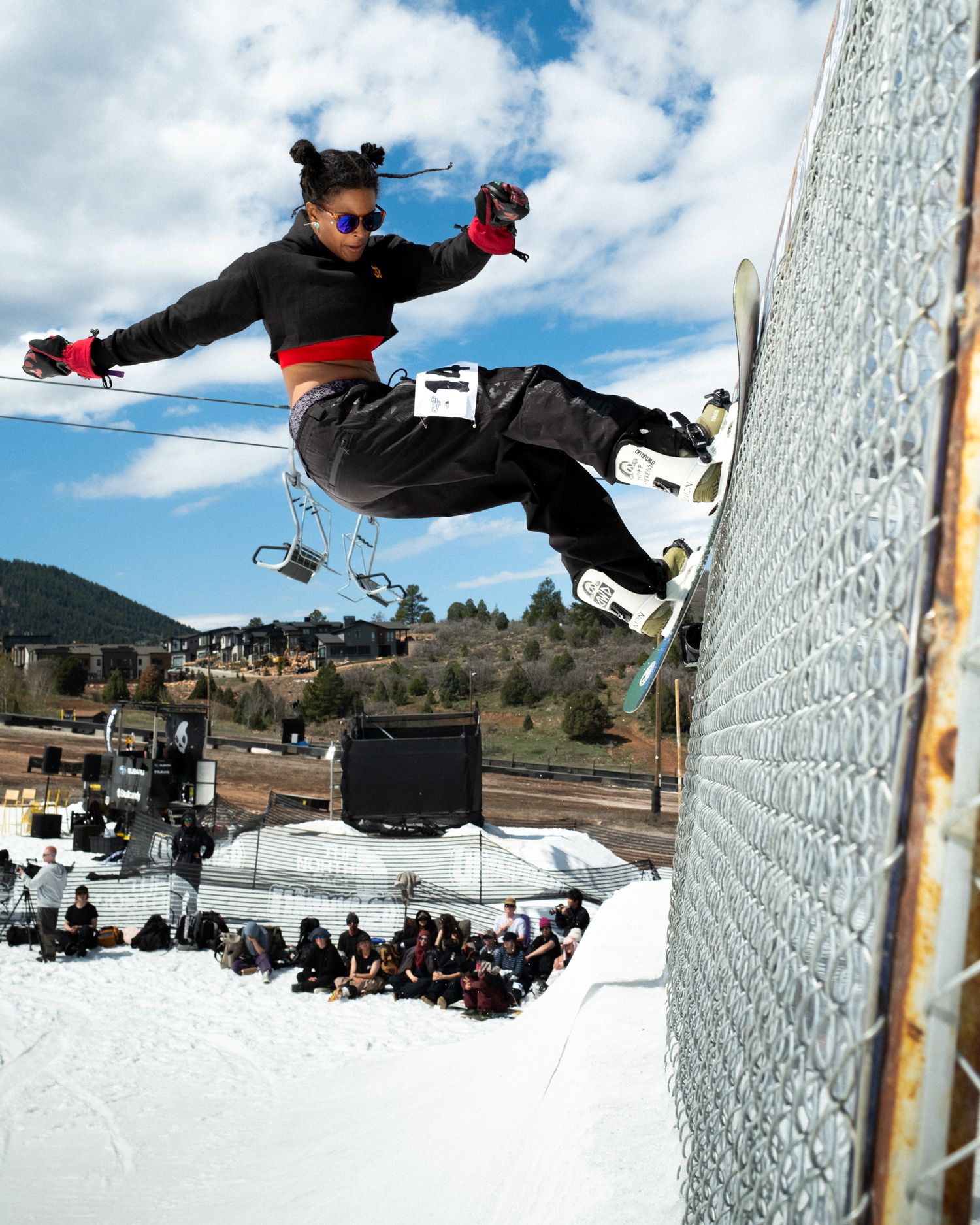 Snowboarder performing a wall ride trick at a snow park with spectators watching under a partly cloudy sky.