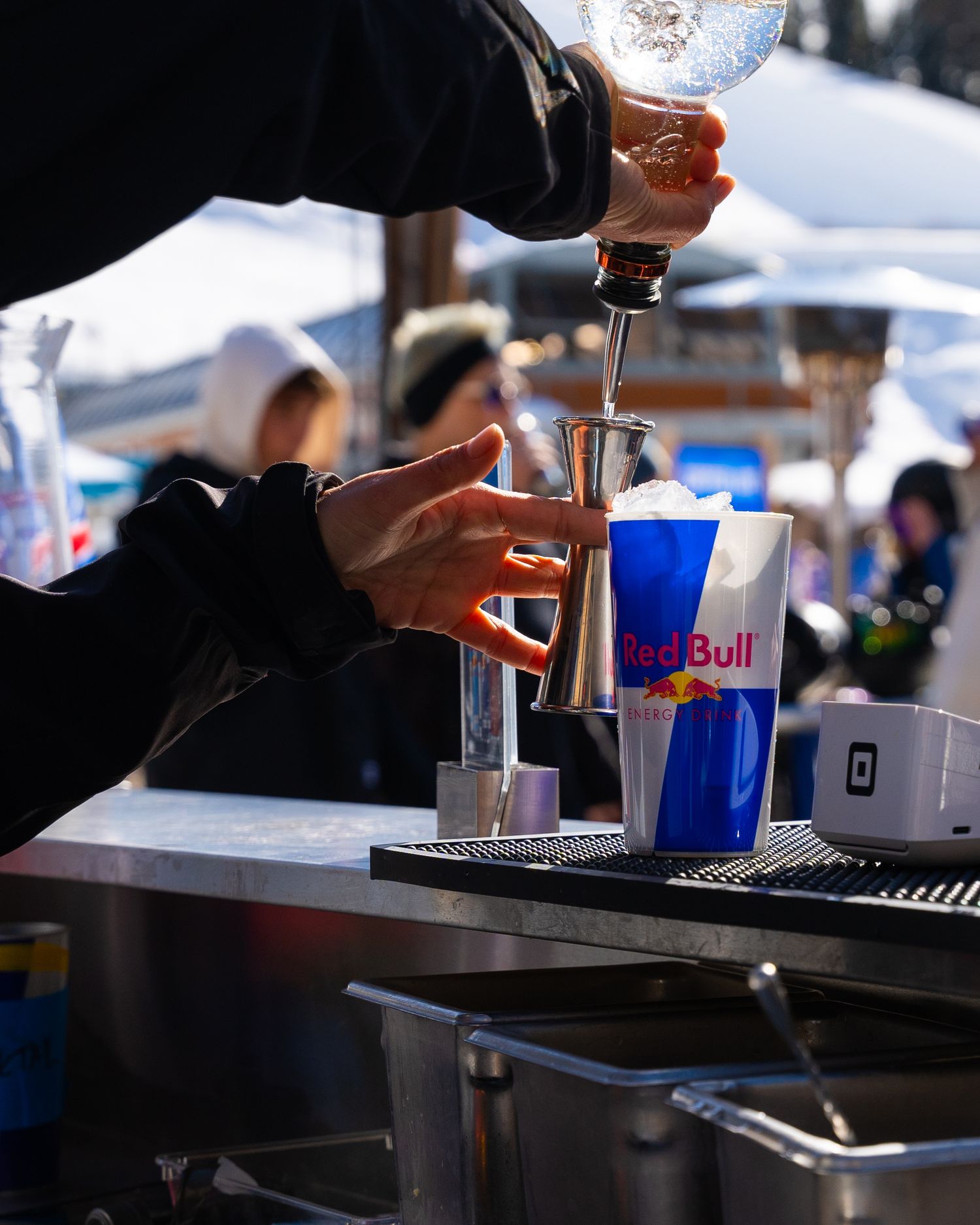 Hands pouring a Red Bull drink into blue branded cup at outdoor winter event.