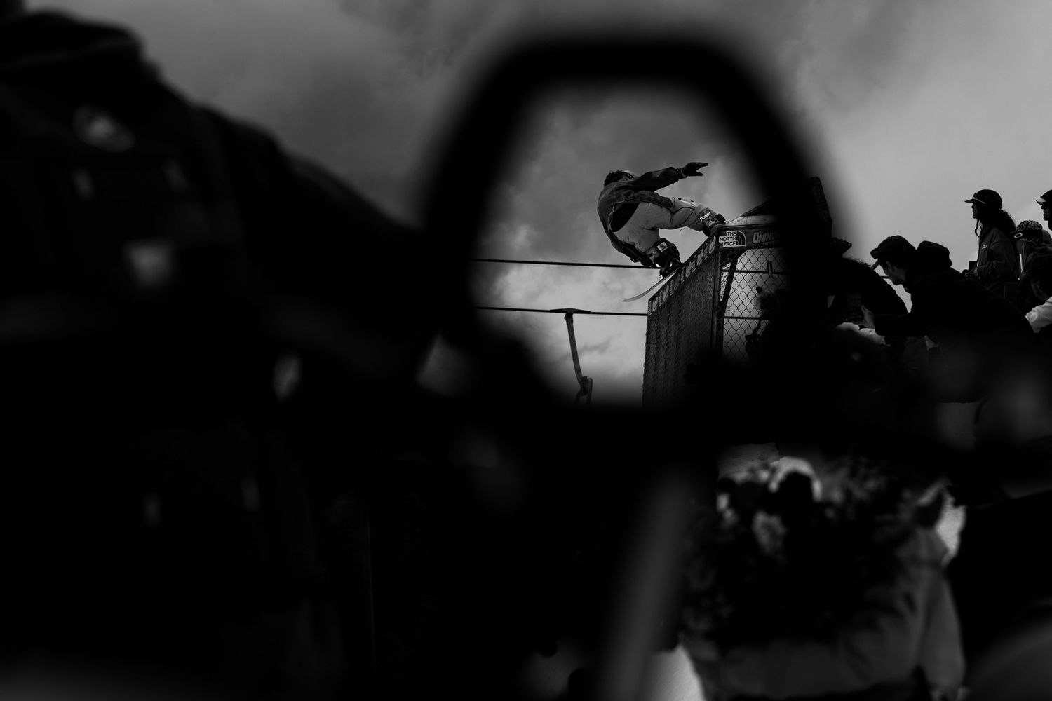 Black and white photo of a skateboarder performing a trick, viewed through a circular frame with crowd in background.