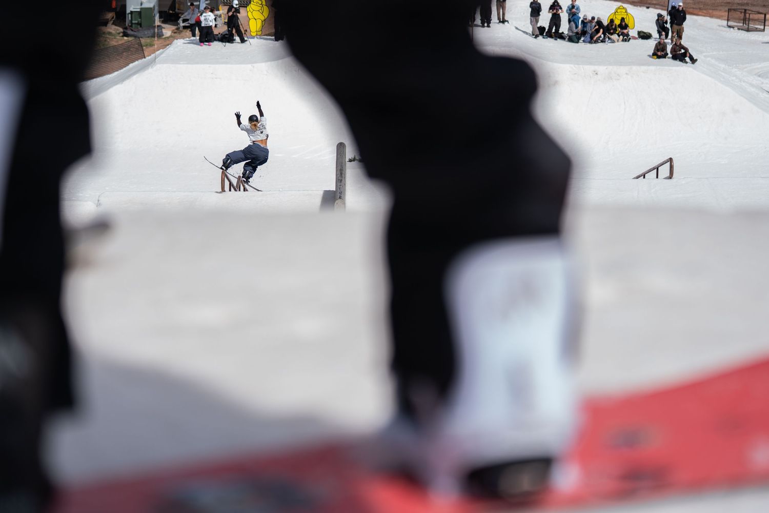 Skateboarder performing a trick at an outdoor skate park while spectators watch in the background on a sunny day.