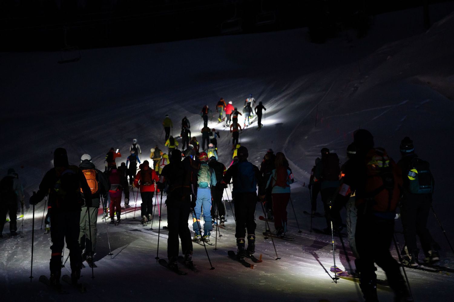 Silhouettes of skiers with headlamps navigating a nighttime slope under moonlit sky.