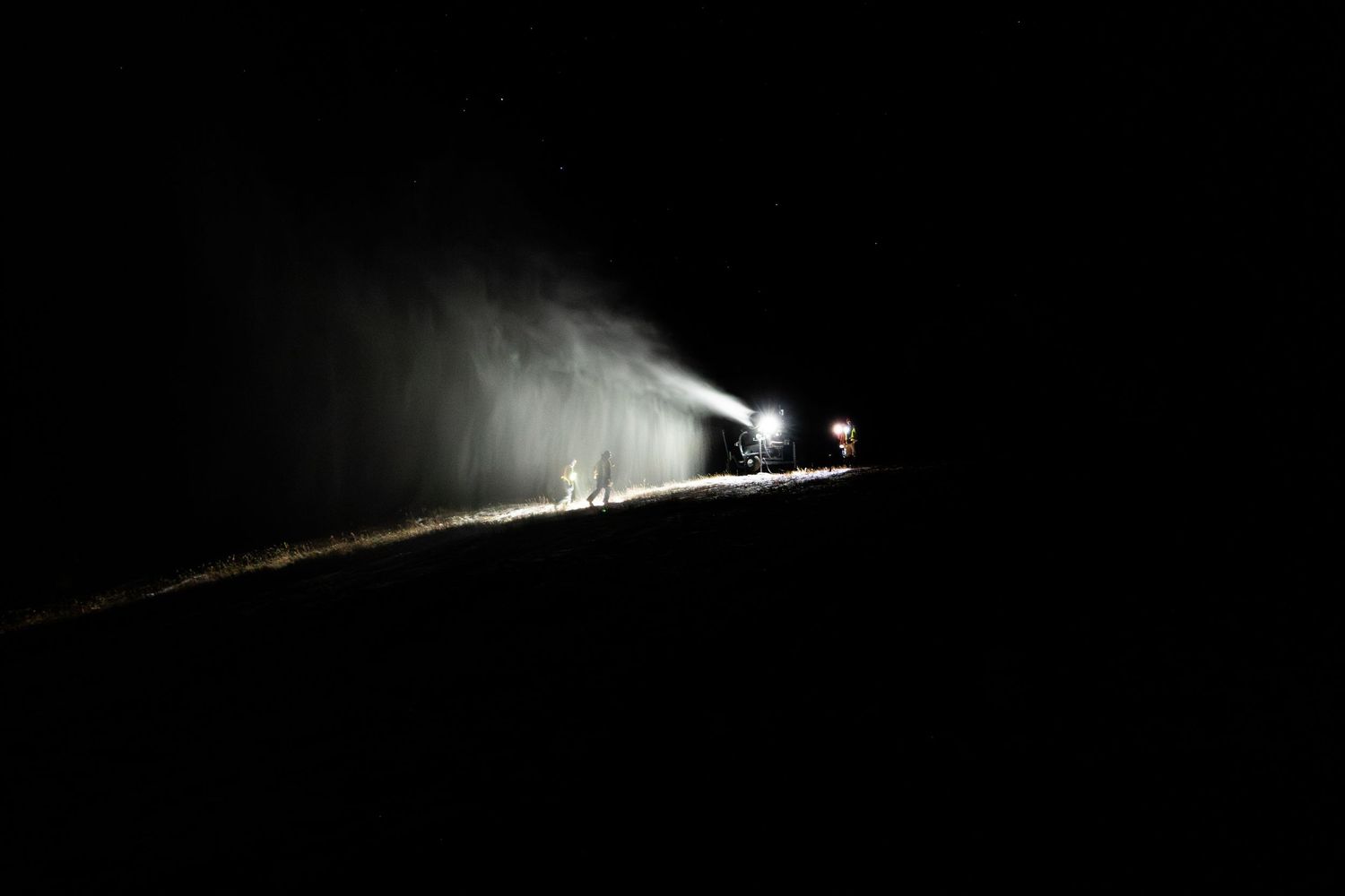 Nighttime snowmaking operations creating illuminated snow spray on dark mountain slope.