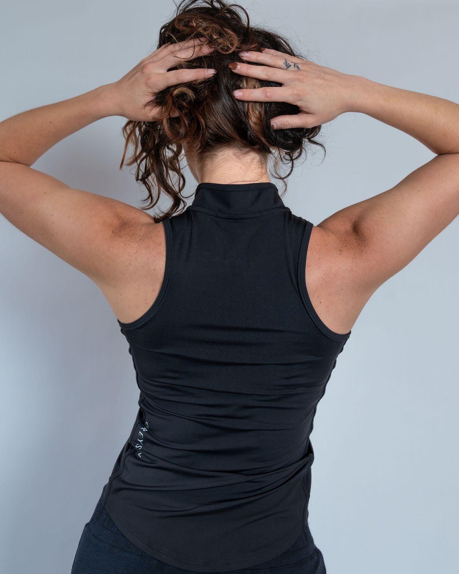Woman wearing a black sleeveless mock neck athletic top, shown from the back with hands raised to her head.