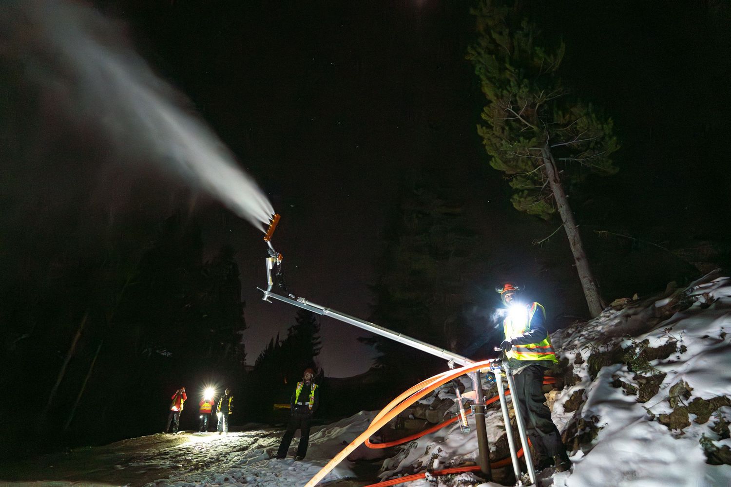 Snow-making machines spray artificial snow at night on ski slopes with workers visible in the distance.