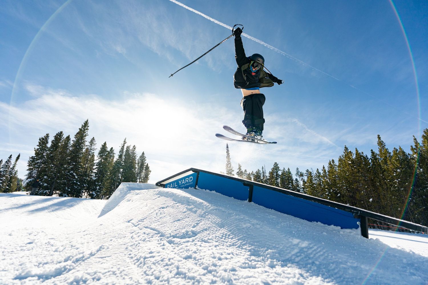 Freestyle skier performing aerial trick off jump in snow park with evergreen trees in background.