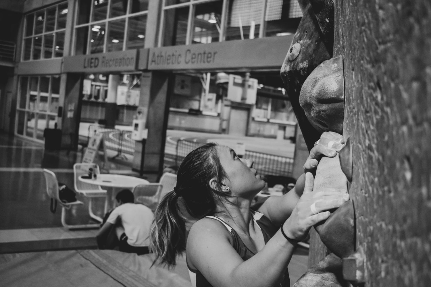Climber navigating holds on an indoor rock climbing wall beside windows.