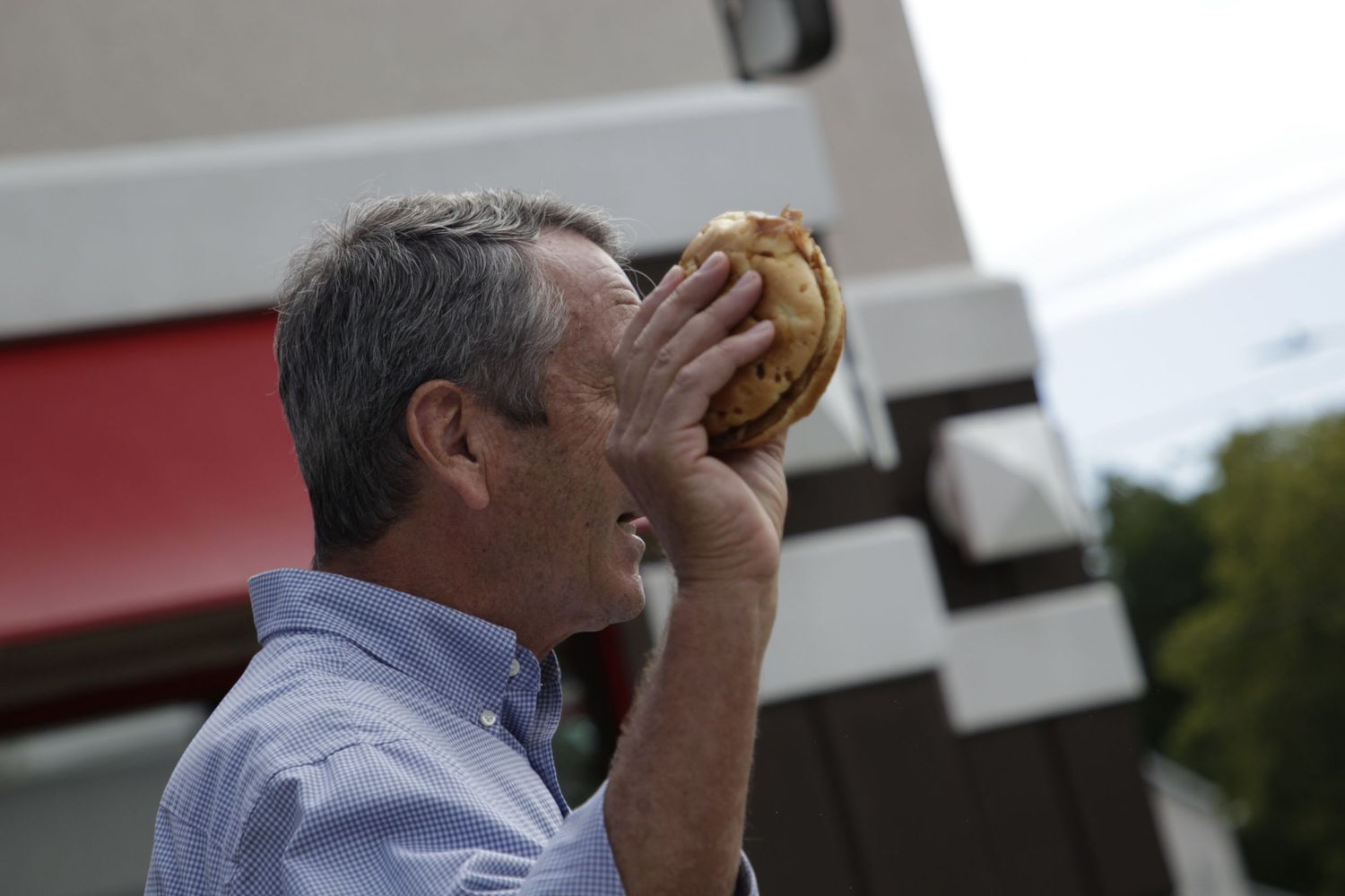 Man in business attire holding a sandwich at an outdoor campaign event.