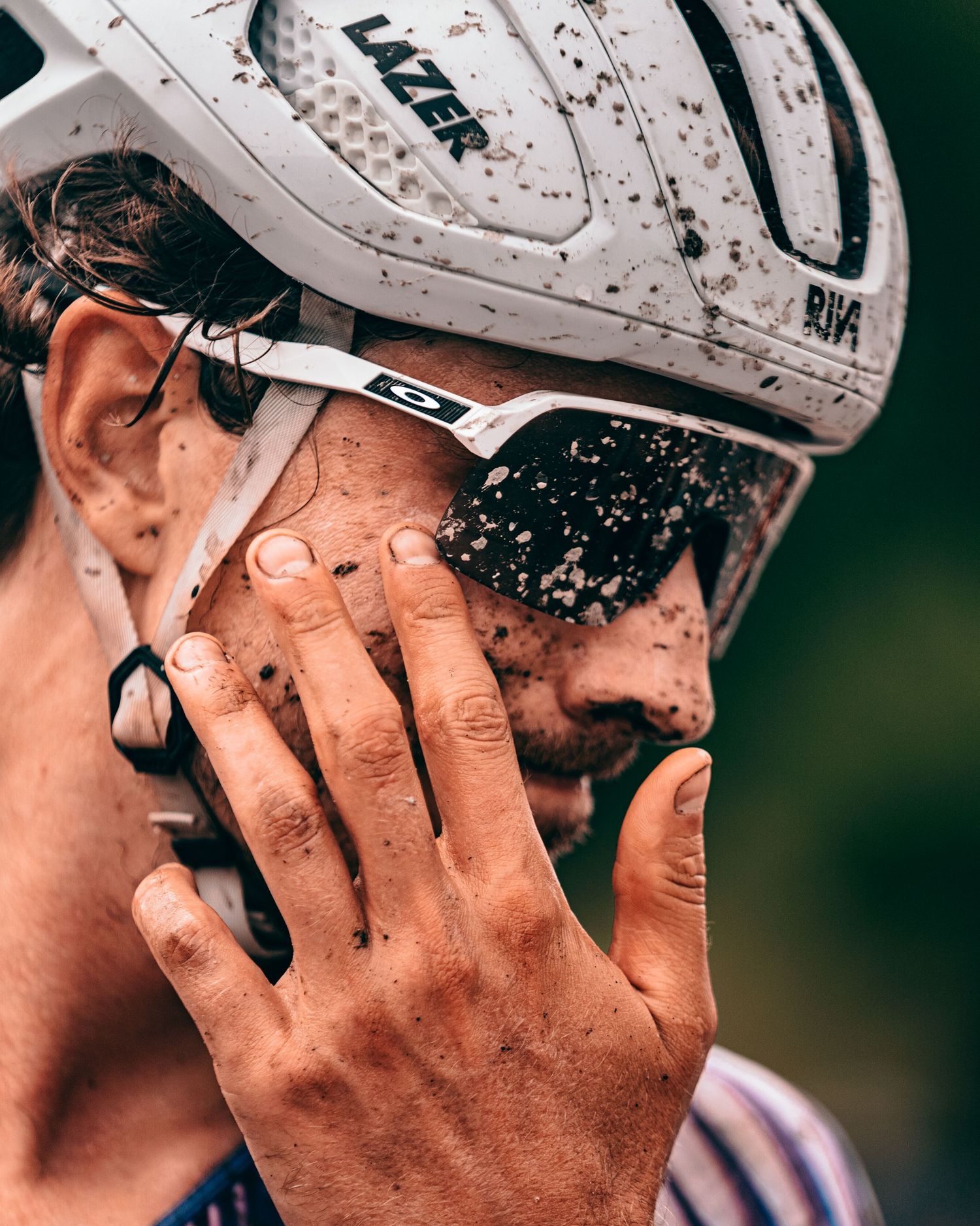 Muddy cyclist wearing a Lazer helmet and Oakley sunglasses wipes dirt from face after a race.