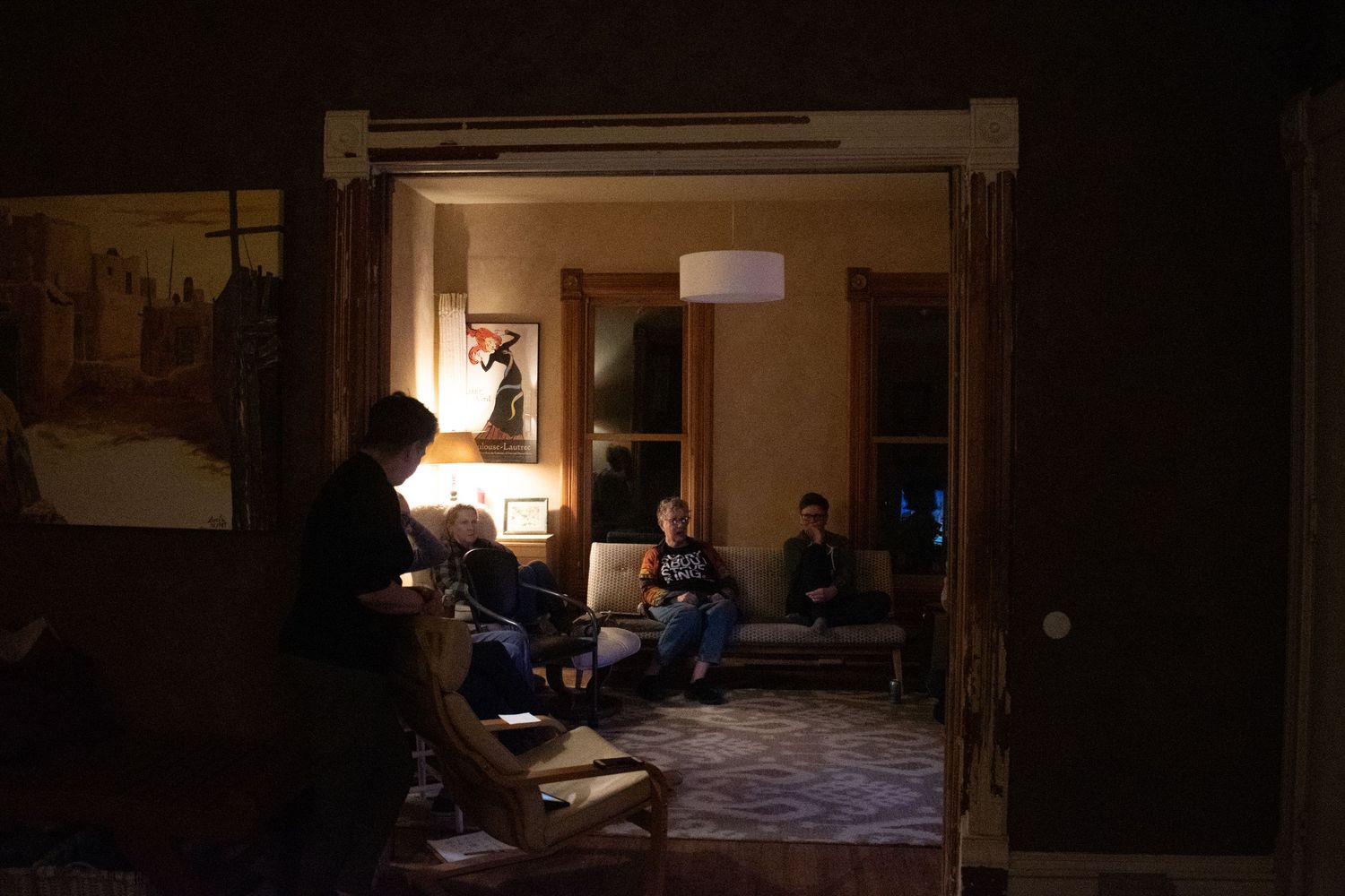 People relax in dimly lit room with warm lamp light casting shadows through doorway in evening atmosphere.
