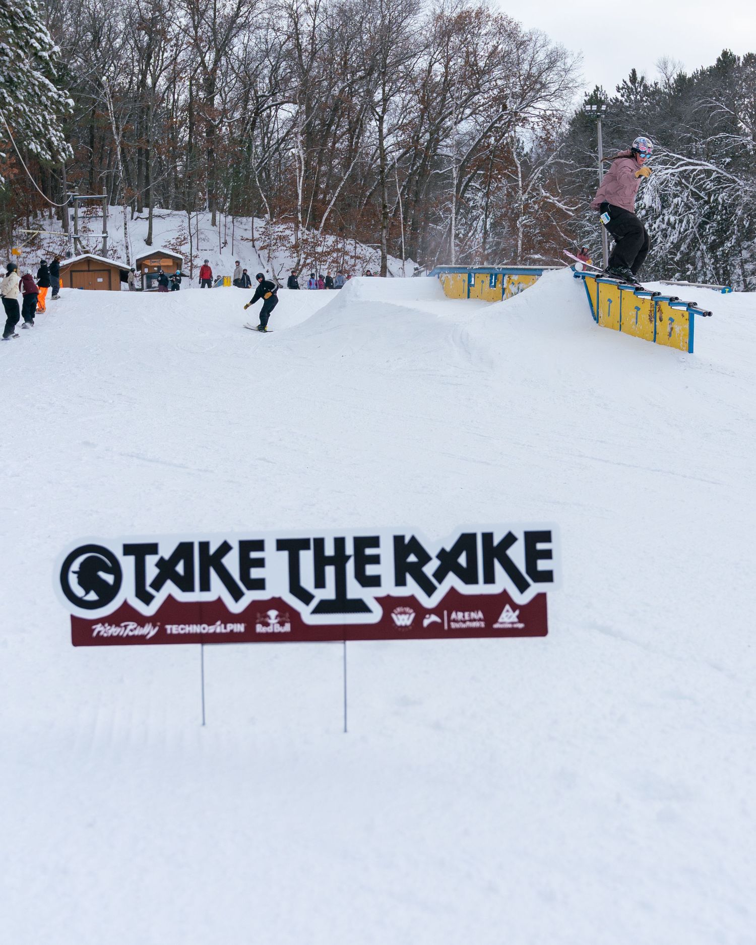Snowboarder catches air at Take The Rake winter terrain park competition event on a snowy day.