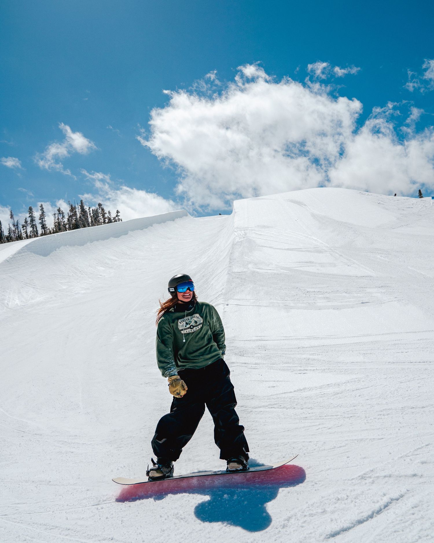 Snowboarder riding down groomed slope under bright blue winter sky.