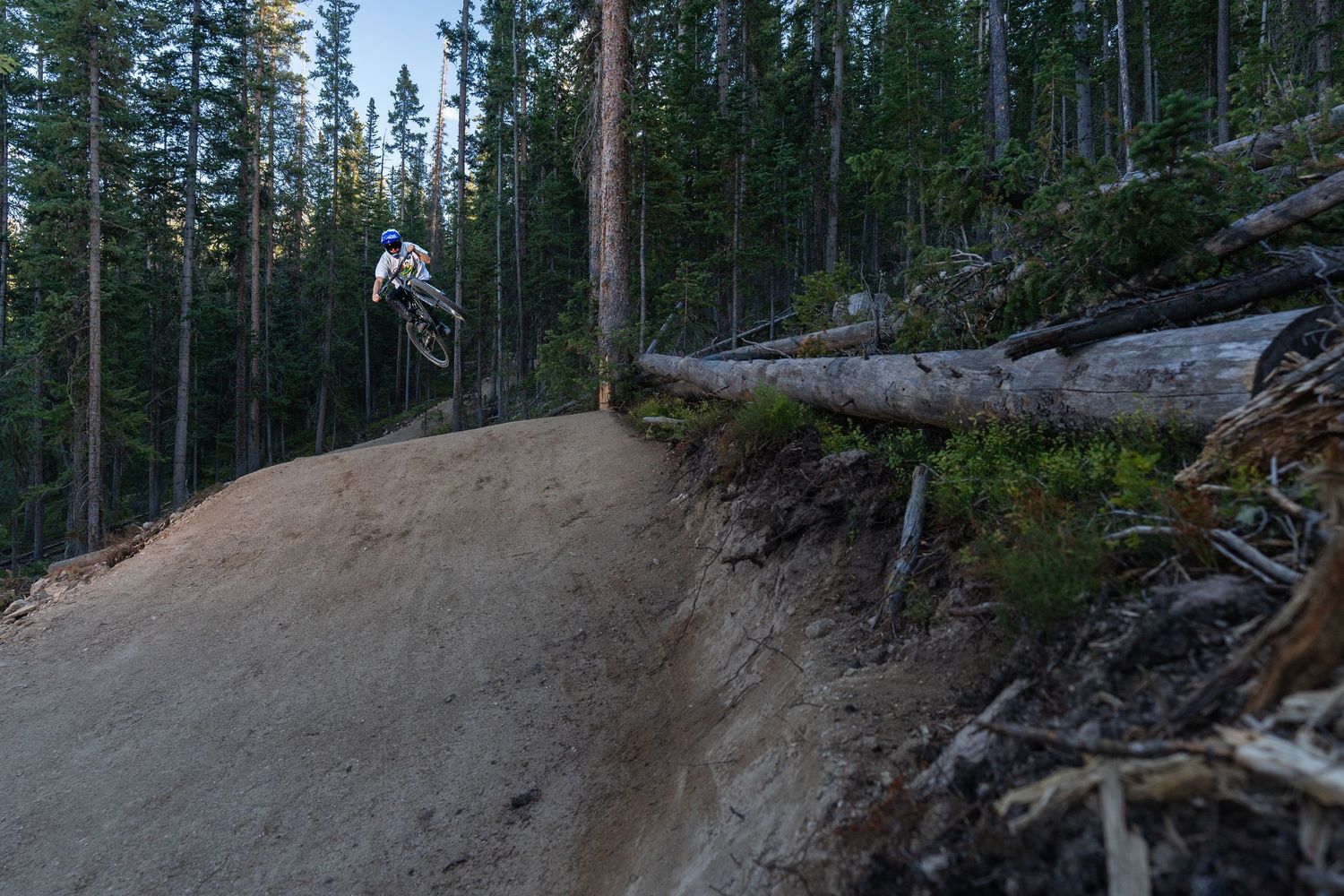 Mountain biker silhouetted while catching air off dirt jump at dusk in forest setting.