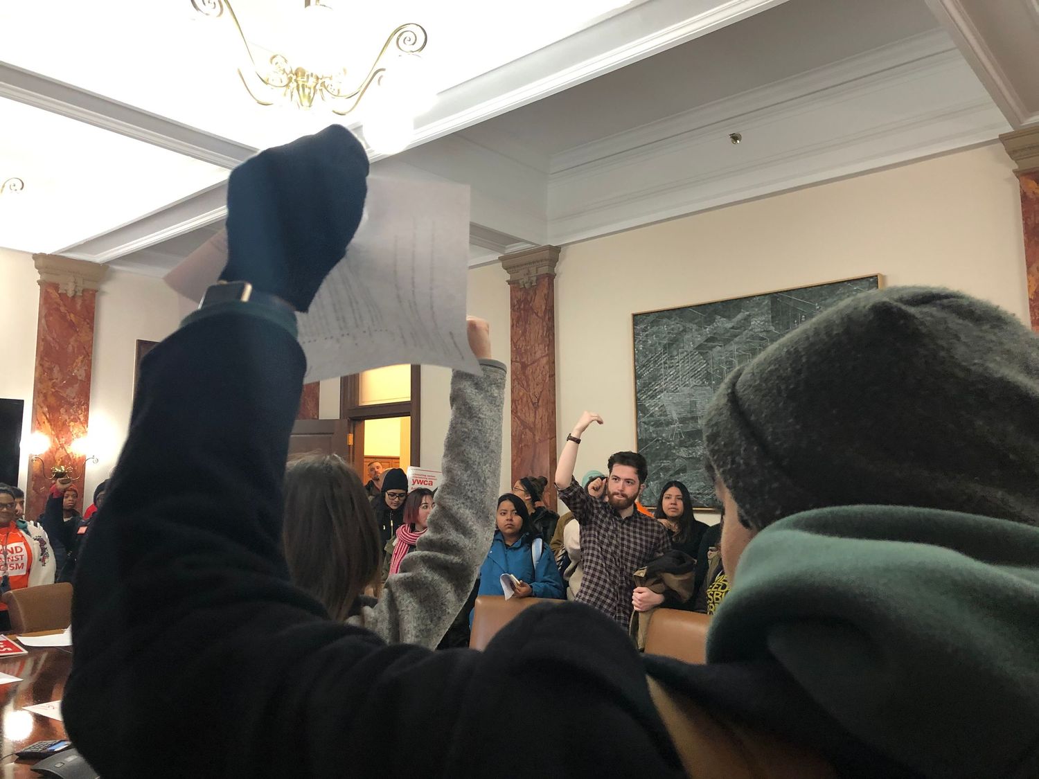 A crowd gathers in an ornate room with raised fists during what appears to be a meeting or rally.