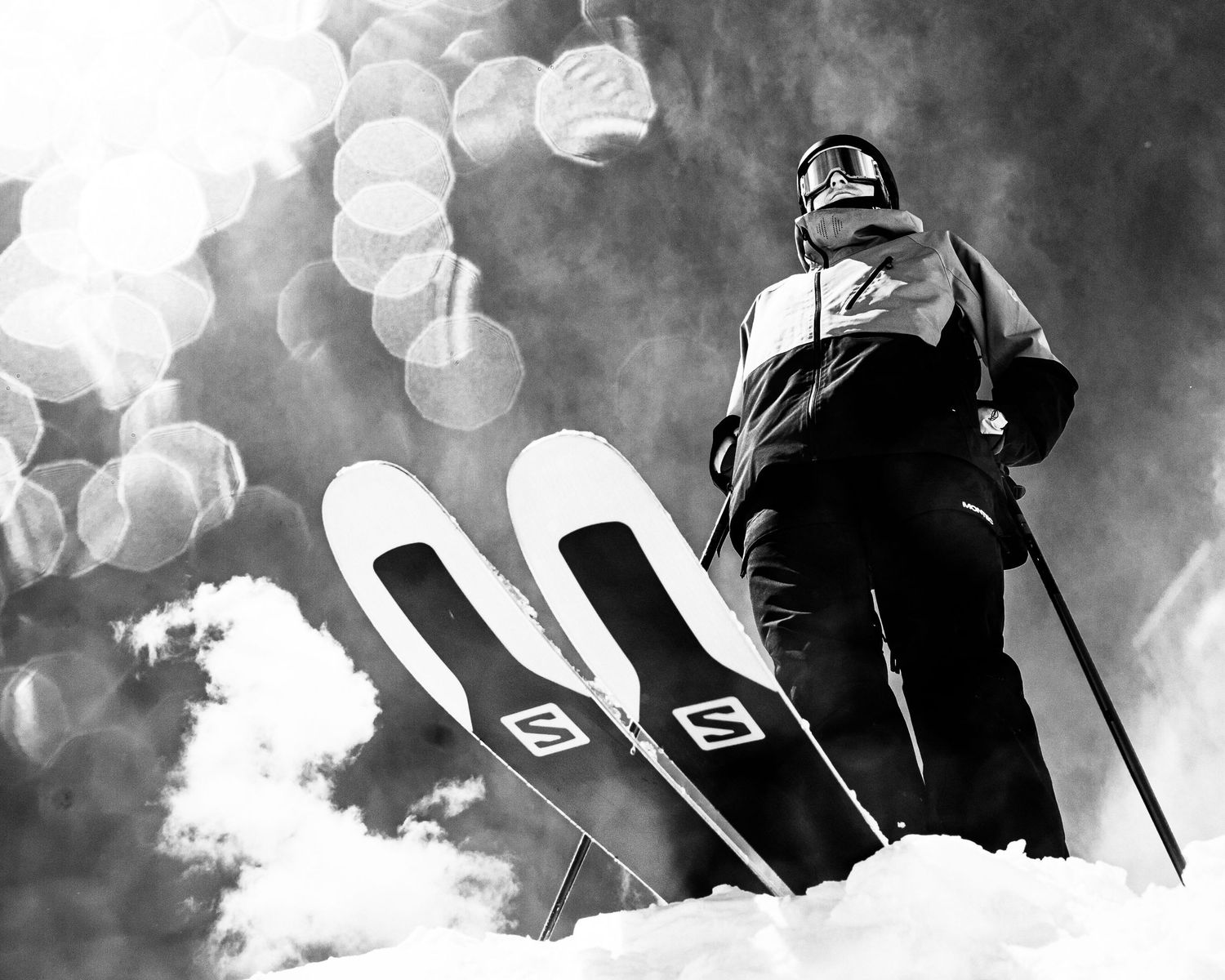 A dramatic black and white shot of a skier with Salomon skis silhouetted against a bokeh-filled winter sky.