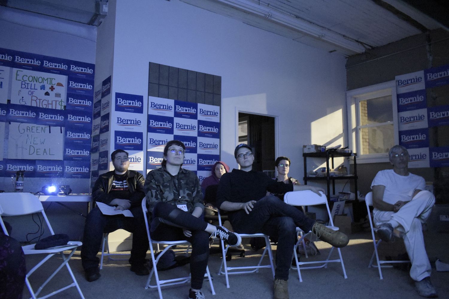 A group sits in folding chairs against a wall with blue and white branded banners in a dimly lit room.