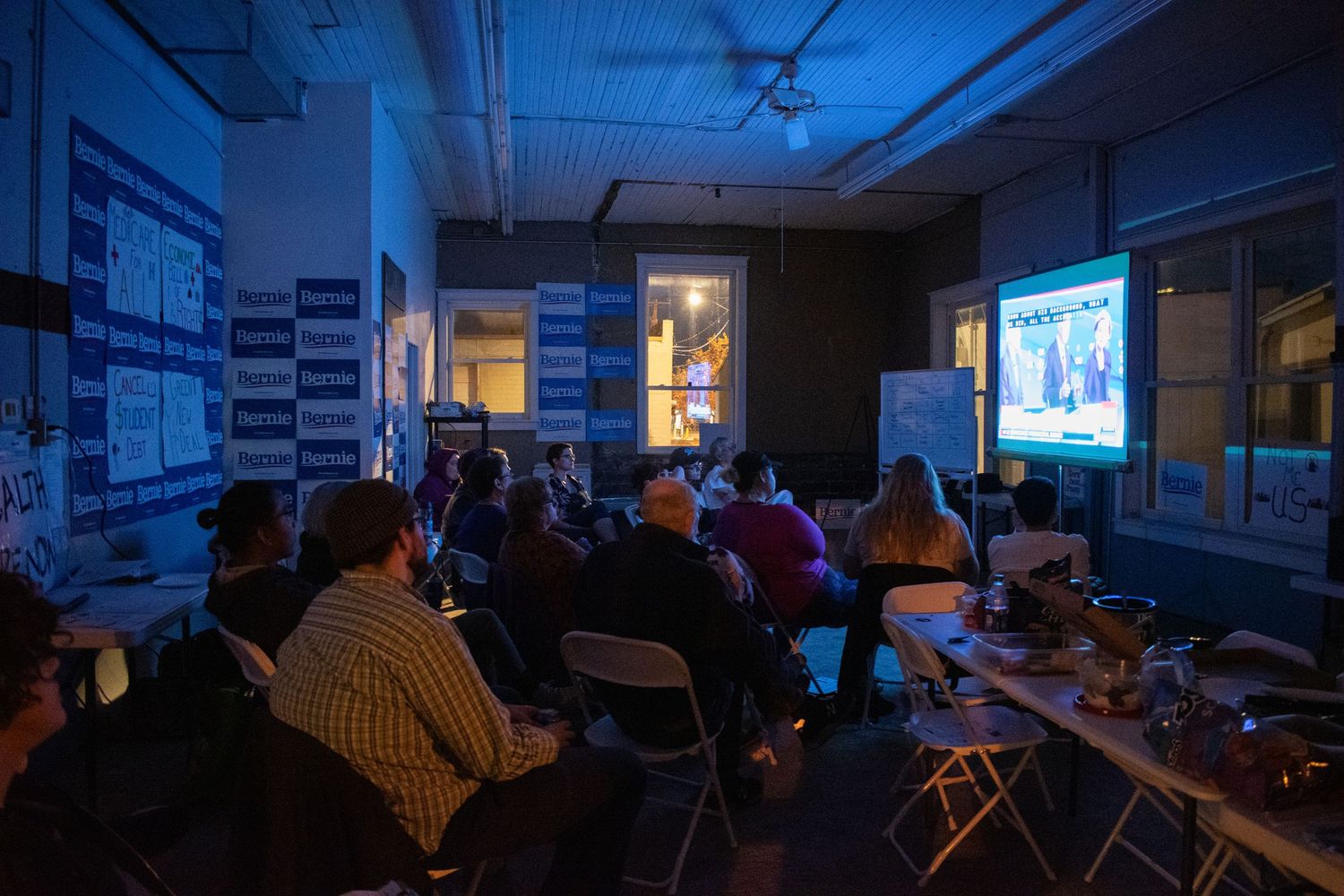 People watching a presentation on a projector screen in a dimly lit room with blue lighting.