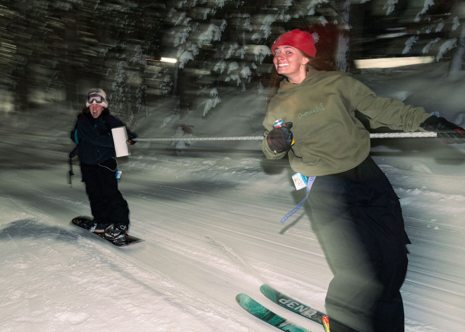 Two skiers enjoying a night ski run on a snowy slope, one wearing a red hat and green jacket leading the way.