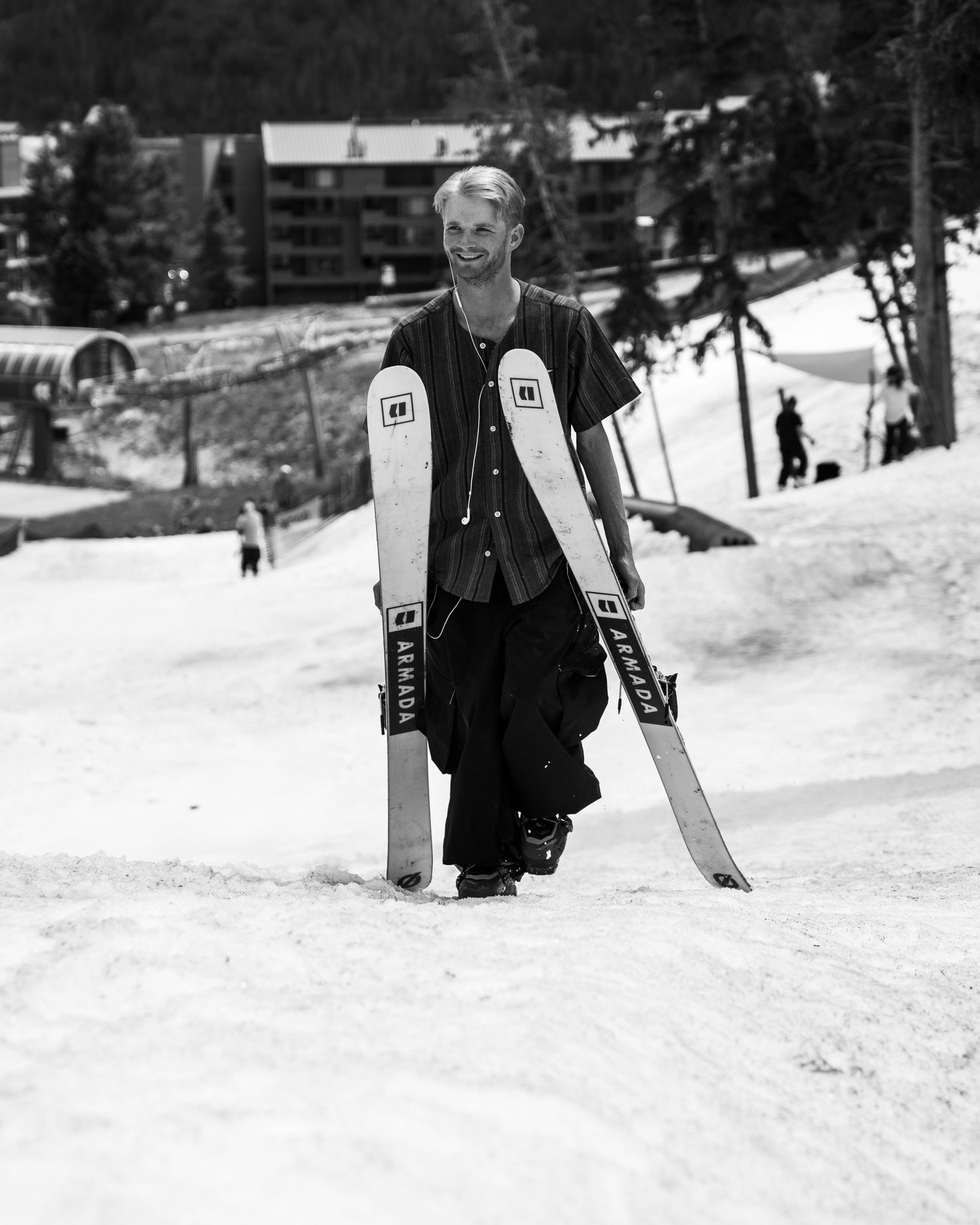 Skier standing on snowy slope holding twin tip skis with lodge buildings in background.