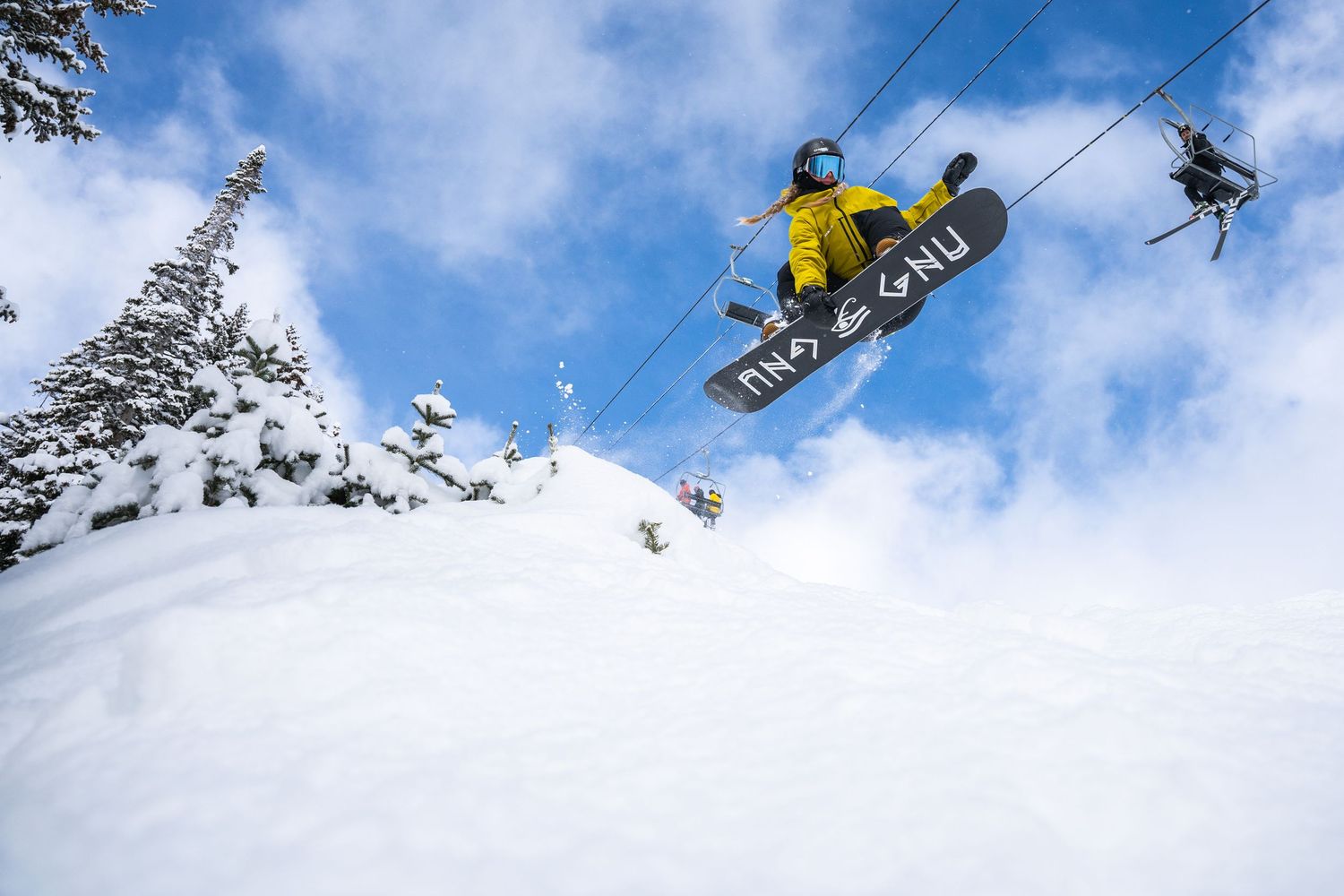 Snowboarder in yellow jacket performs aerial jump against snowy mountain backdrop and blue sky.