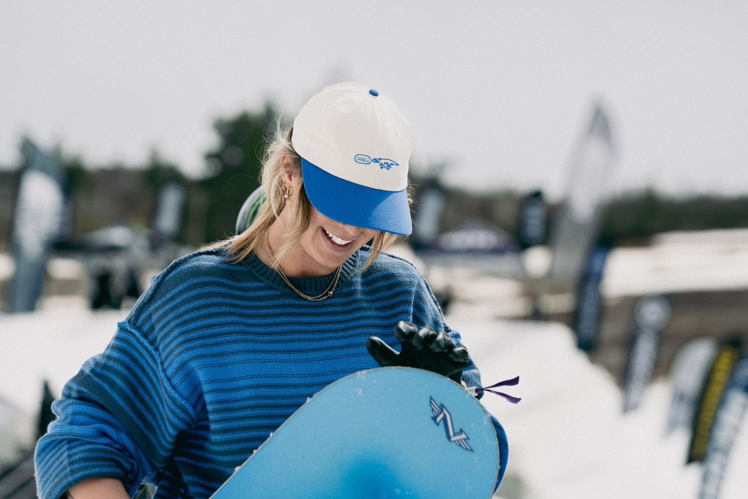 Smiling female snowboarder in blue striped sweater holding a snowboard at an outdoor event.
