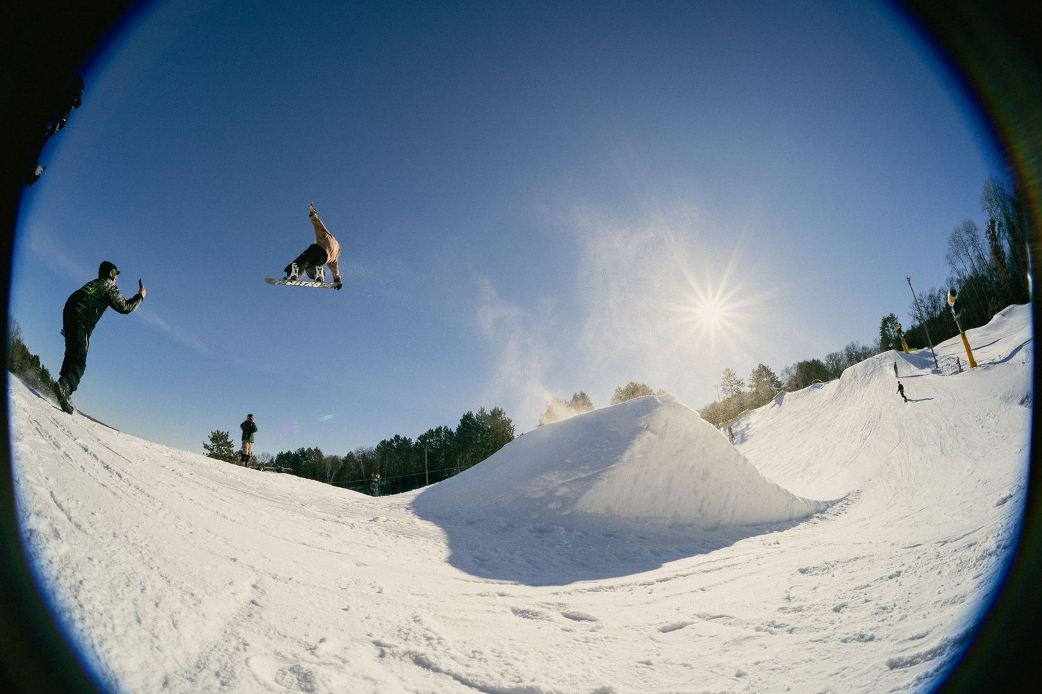 Snowboarder performing aerial trick off snow jump at terrain park on sunny day, captured with fisheye lens.