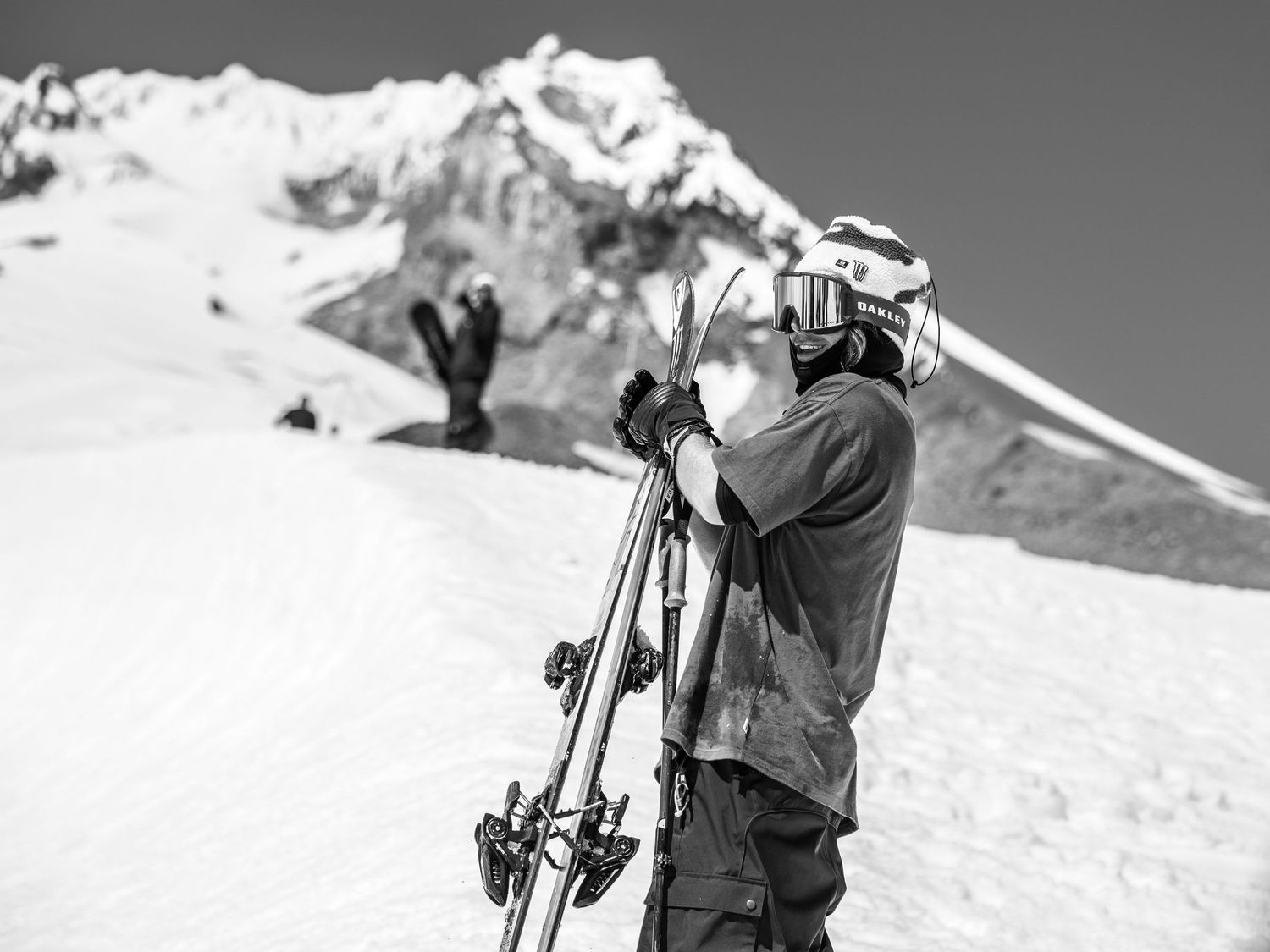 Black and white photo of skier carrying equipment with snow-capped mountain in background.
