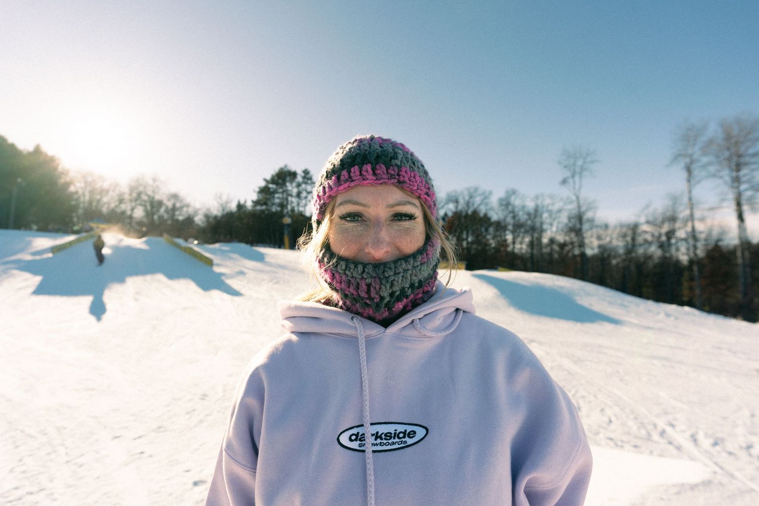 Person wearing a colorful knit balaclava and white hoodie standing in a snowy ski park on a sunny winter day.