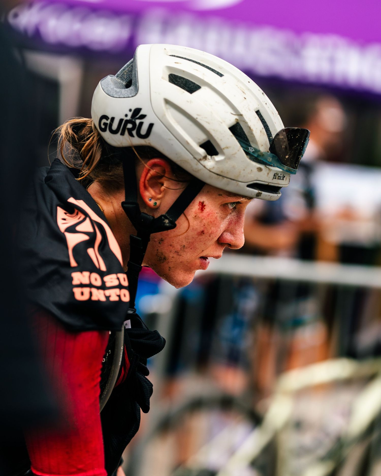 Female cyclist wearing a white Guru helmet with mud on her face competing in an intense cycling race event.