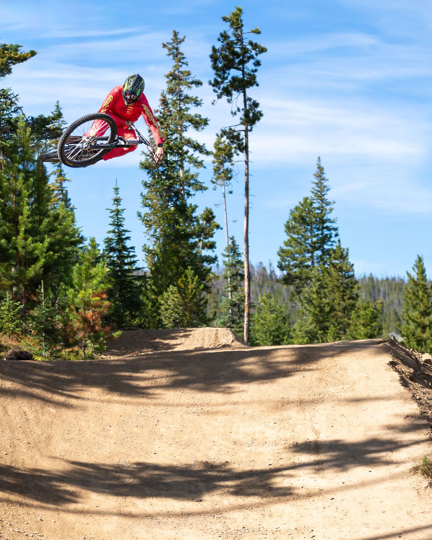 Mountain biker performs impressive jump on curved dirt trail in forest setting.